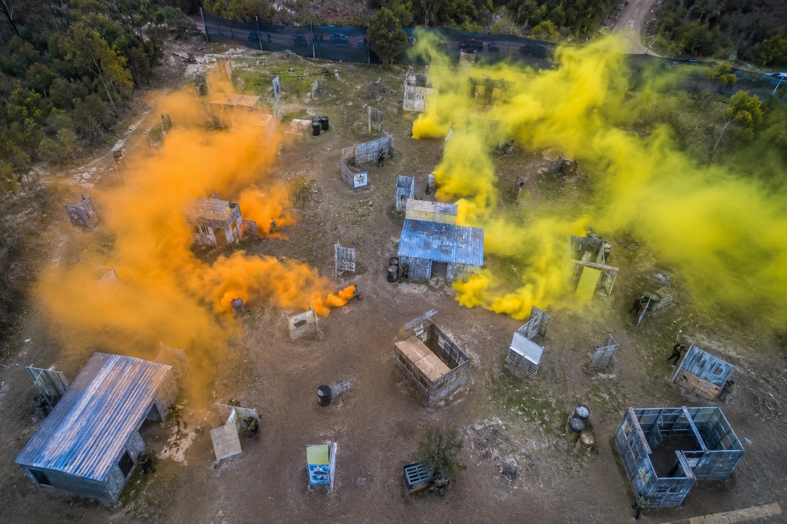 Aerial drone shot of a large outdoor paintball arena with smoke grenades and players crouched behind bunkers